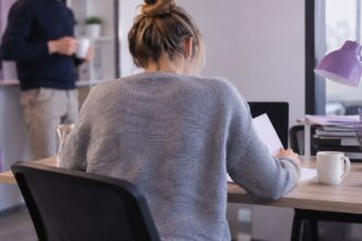 Personne assise de dos à un bureau, coudes posés, tasse blanche, silhouette floue en arrière-plan.