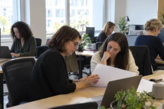 Plusieurs femmes de tous âges travaillent ensemble dans un open space moderne et lumineux, certaines devant leur ordinateur, deux d'entre elles discutent un dossier