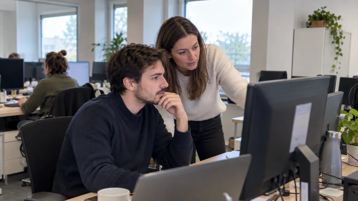 Un nouveau collaborateur travaille à son poste dans un open space moderne pendant que sa tutrice, penchée vers lui, lui explique un point à l'écran vu de biais