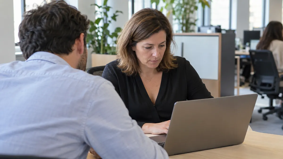Une DRH consulte un dossier numérique sur son ordinateur portable à un bureau moderne, un collègue de dos près d'elle, open space clair en arrière-plan, lumière naturelle
